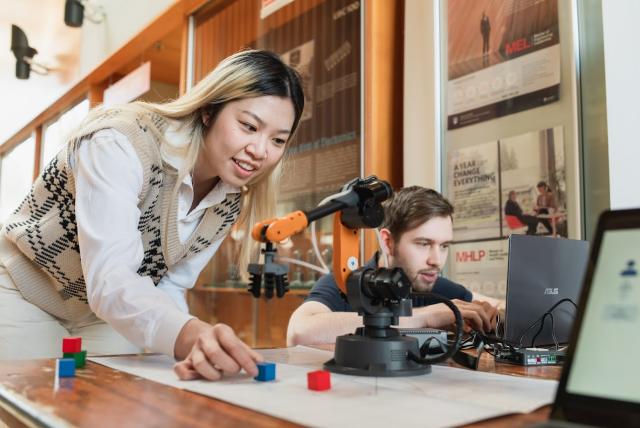 Two people working on a robotic arm and colorful blocks on a table, one using a laptop.