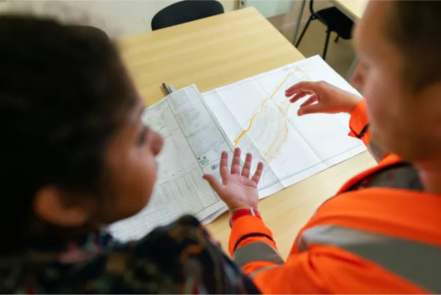 Two people discussing a map spread out on a table, one of them wearing an orange safety vest.