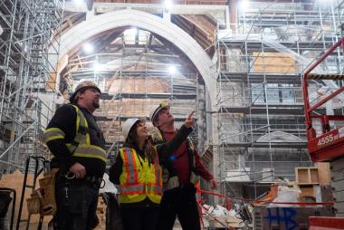 Three construction workers in safety gear discussing inside a large building under renovation.