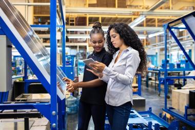 Two women in a manufacturing facility examining a solar panel and using a tablet.