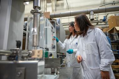 Two women in lab coats operating a machine in an industrial lab setting.