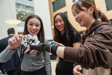 Three women smiling and examining a mechanical hand device.