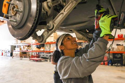 Mansi Tandon working at Rivian. She standing under a raised vehicle, looking upward with a drill pointing up to the underside of the vehicle.